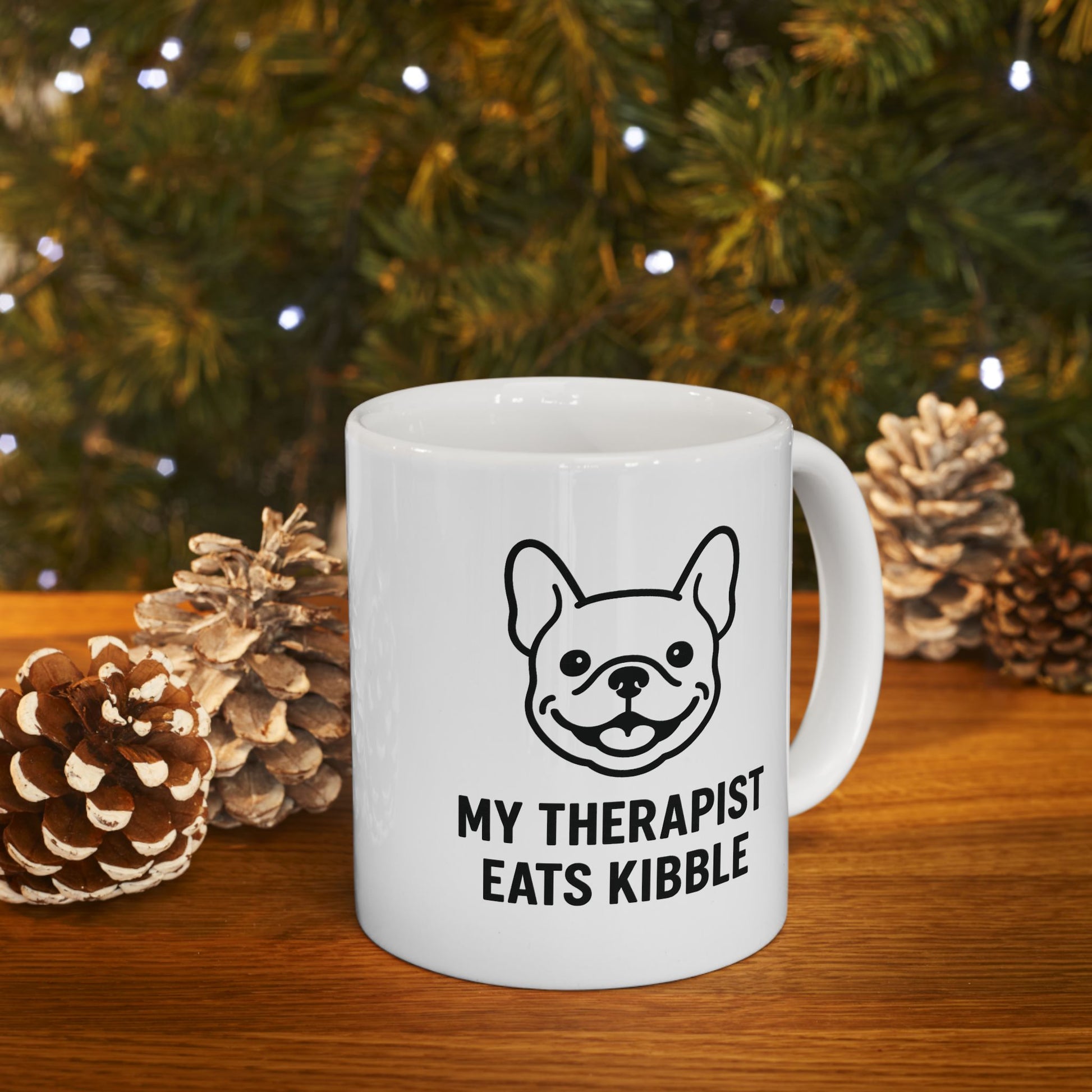 White mug with a dog illustration and text in front of a Christmas tree and pinecones.